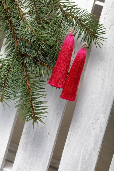 Pink tassel earrings hang on a spruce branch. Against the background of a white wooden box.