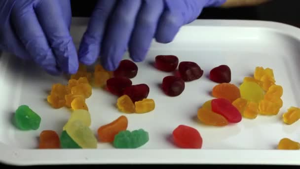 Gelee-Bonbons in verschiedenen Formen und Farben sind auf einem Tablett verstreut. Ein Mann in Gummihandschuhen sortiert sie in Sets. Internationaler Süßigkeitstag. — Stockvideo