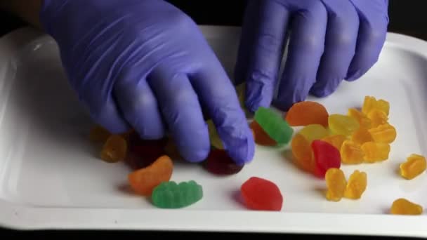 Een man met rubberen handschoenen sorteert snoepjes. Sweets van verschillende vormen en kleuren worden verspreid over een dienblad. Internationale Snoepdag. — Stockvideo