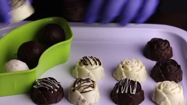 Egy gumikesztyűs férfi a tálcáról csokoládét visz egy konténerbe. Mázas édességek fehér és fekete csokoládéban. A cukorka nemzetközi napja. — Stock videók