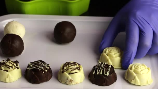 Un uomo in guanti di gomma attraversa caramelle al cioccolato da un contenitore su un vassoio. Dolci glassati al cioccolato bianco e nero. Giornata internazionale della caramella . — Video Stock