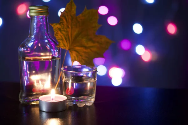 Candela accesa con un drink sullo sfondo con luci colorate. Nelle vicinanze si trovano un bicchiere e una bottiglia di bevanda. Tirato fuori foglie di acero di autunno. Accessori vacanze al crepuscolo . — Foto Stock