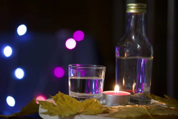 Candela accesa con un drink sullo sfondo con luci colorate. Nelle vicinanze si trovano un bicchiere e una bottiglia di bevanda. Tirato fuori foglie di acero di autunno. Accessori vacanze al crepuscolo . — Foto Stock