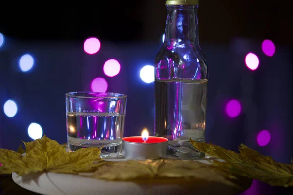 Candela accesa con un drink sullo sfondo con luci colorate. Nelle vicinanze si trovano un bicchiere e una bottiglia di bevanda. Tirato fuori foglie di acero di autunno. Accessori vacanze al crepuscolo . — Foto Stock