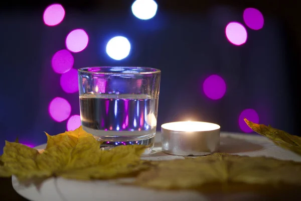 Vela ardente e um copo com uma bebida no fundo com luzes coloridas. Perto estão as folhas de bordo do outono. Acessórios de férias ao entardecer . — Fotografia de Stock