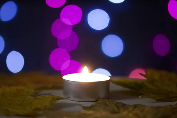 Vela ardiendo sobre un fondo con luces de colores. Cerca están las hojas de arce de otoño. Accesorios de vacaciones al atardecer . — Foto de Stock