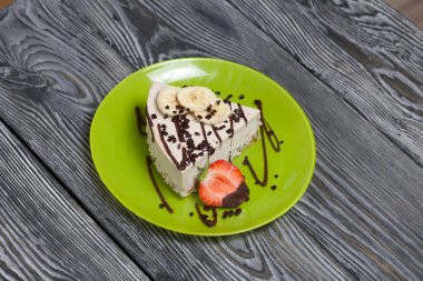 Tabakta peynirli kek. Çilek, muz ve çikolatayla süslenmiş. Siyah beyaza boyanmış çam ağacından tahtalara.