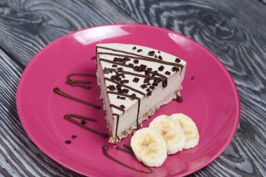 Tabakta peynirli kek. Muz ve çikolatayla süslenmiş. Siyah beyaza boyanmış çam ağacından tahtalara.