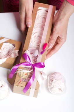 Kağıt ambalajda marşmelov ve kadın eli. Kurdelelerle ve kurumuş çiçeklerle süslenmiş. Gül şeklinde marşmelovlar. Beyaz arkaplanda.