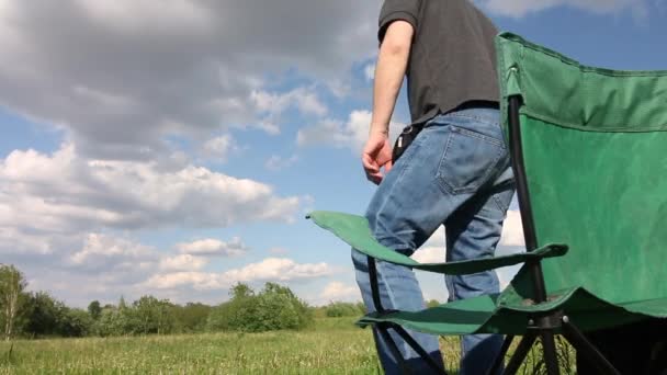 Un hombre se sienta en una silla de camping. Pradera con hierba verde. Cielo azul con nubes . — Vídeo de stock