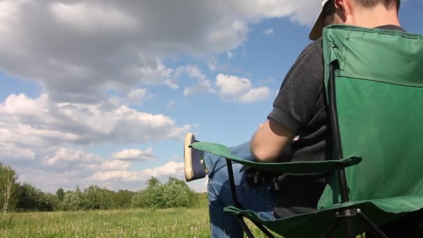 男はキャンプチェアに座って、電話で彼の前の風景を撮影します。緑の芝生の牧草地。雲のある青空. — ストック動画