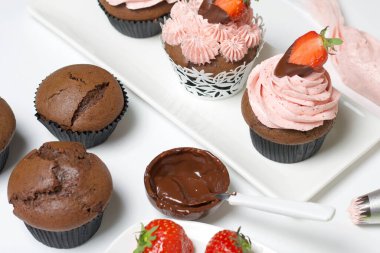 Çilekli kremalı çikolatalı kekler. Yarım çilek ve çikolatayla süslenmiş. Kekler için gerekli olan boş ve malzemelerin yanında..