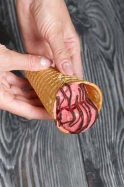 Kadın elinde waffle külahında çilekli dondurma tutuyor. Çikolatayla süslenmiş. Siyah beyaz boyanmış çam ağaçlarının arka planında.