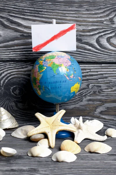 Starfish and shells are scattered on pine boards painted in black and white. There is a globe and a pointer above it with a field for text. The field is crossed out by a red line. Holiday at sea 2020.
