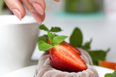 Çilekli ve taçyapraklı çikolatalı şekerleme. Kadın tatlıyı nane ve çilekle süslüyor. Masanın beyaz yüzeyinde.