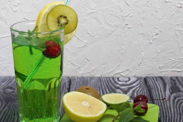 在杯子里放绿色柠檬水 它漂浮着樱桃和薄荷叶 用一片柠檬和猕猴桃装饰 用稻草当鸡尾酒 切菜板上的水果是鸡尾酒的水果 — 图库照片