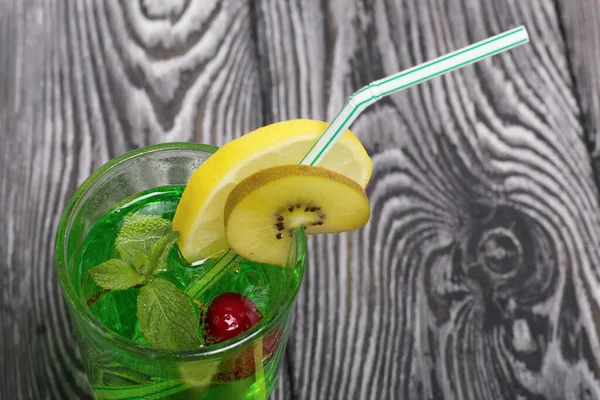 Green lemonade in a glass. It floats cherry and mint leaves. Garnished with a slice of lemon and kiwi. With a straw for a cocktail.
