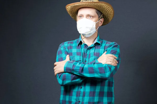 Farmer with a protective mask on his face. There is a straw hat on his head. Arms crossed on the chest. Shot on a gray background.