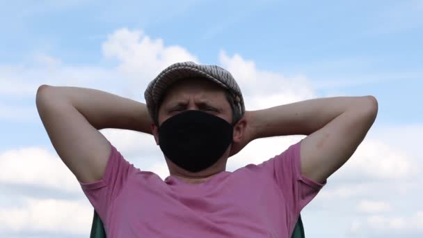 Un hombre con una máscara protectora y gorra se sienta en una silla de camping. En el fondo del cielo azul. Pone las manos detrás de la cabeza. Recreación al aire libre en autoaislamiento durante una pandemia . — Vídeos de Stock