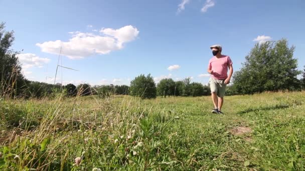 Un hombre con una máscara protectora y gorra está caminando en un prado verde. Un paseo al aire libre aislado durante una pandemia . — Vídeos de Stock