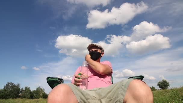 Un hombre con una máscara protectora y gorra se sienta en una silla de camping. Se quita la máscara, bebe agua de una botella. Recreación al aire libre en autoaislamiento durante una pandemia . — Vídeo de stock