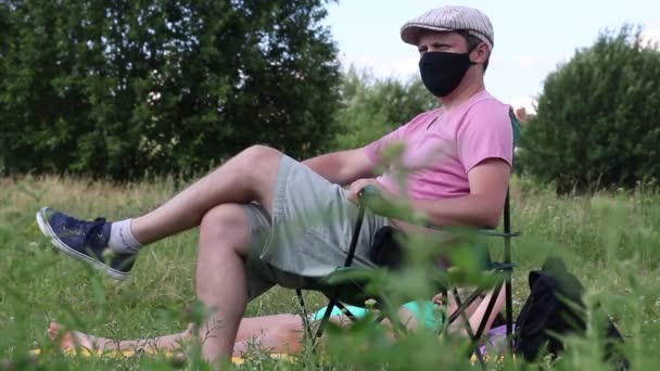 En man i skyddsmask och mössa sitter i en campingstol. Upp och hoppa. En kvinna solar på marken. Friluftsliv i självisolering under en pandemi. — Stockvideo