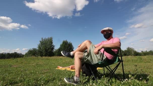 Un uomo con una maschera protettiva e un berretto si siede su una sedia da campeggio. Una donna sta prendendo il sole a terra. Attività ricreative all'aperto in auto-isolamento durante una pandemia — Video Stock