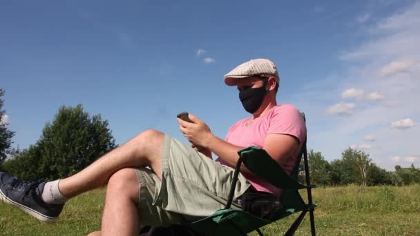 Koruyucu maskeli ve şapkalı bir adam kamp koltuğunda oturuyor. Telefonla konuşuyorum. Bir kadın yerde güneş banyosu yapıyor. Bir salgın sırasında kendini izole ederek dışarıda dinlenmek. — Stok video