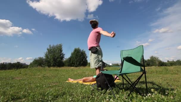 A man in a protective mask and cap spreads his hands and sits in a camping chair. A woman is sunbathing on the ground. Outdoor recreation in self-isolation during a pandemic. — Stock Video