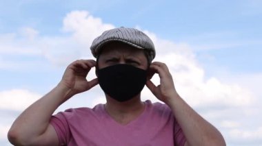 Şapkalı bir adam kamp koltuğunda oturur. Yüz maskesi takıyordu. Mavi gökyüzünün arka planına karşı. Bir salgın sırasında kendini izole ederek dışarıda dinlenmek..