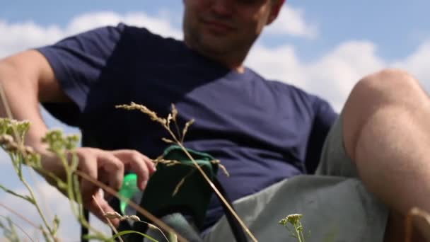 Un hombre se sienta en una silla de camping en un prado verde. Bebe agua de una botella. Relajado, devuelve la cabeza. Salí a tomar aire fresco en el parque de la ciudad. Caminar y descansar durante la epidemia — Vídeos de Stock