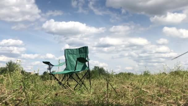 Egy kempingszék áll a réten a zöld fű között. Bolyhos felhők vannak a kék égen. A serpenyőből eltávolítva. — Stock videók