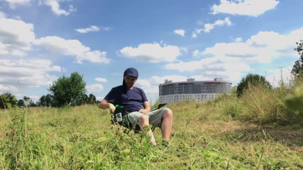 Een man zit in een campingstoel op een groene weide. Hij doet het masker af en drinkt water uit de fles. Wandelen en rusten tijdens de epidemie en zelfisolatie. Algemeen plan. — Stockvideo