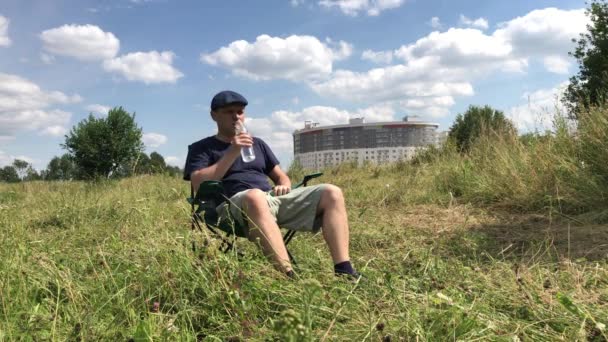 Un hombre se sienta en una silla de camping en un prado verde. Bebe agua de una botella. Humedece el cabello con agua. Caminar y descansar durante la epidemia y el autoaislamiento. Plan global . — Vídeo de stock