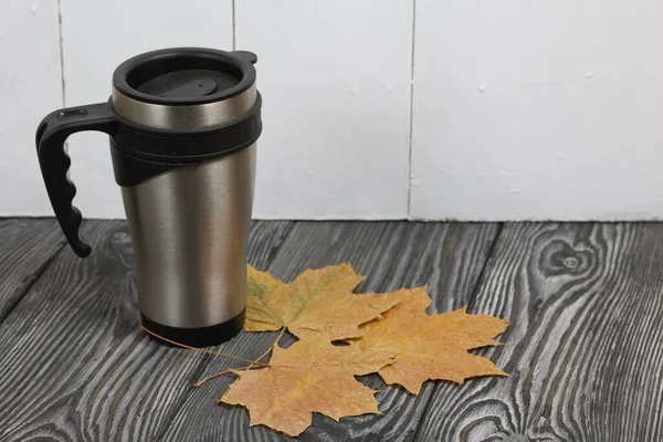 Thermo mug on a pine board surface. Nearby are dried maple leaves.