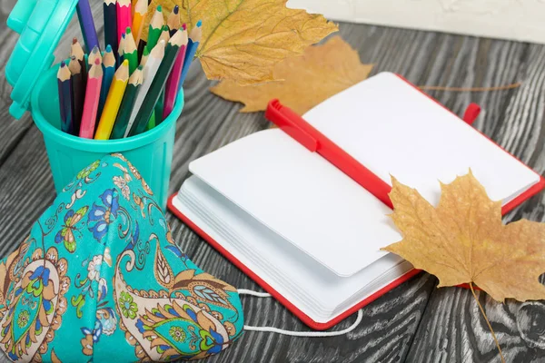 A pencil case in the form of a trash can. It contains colored pencils. Nearby is a notepad and a protective mask for the pandemic. Dried maple leaves are added to the compositions.