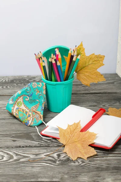 A pencil case in the form of a trash can. It contains colored pencils. Nearby is a notepad and a protective mask for the pandemic. Dried maple leaves are added to the compositions.