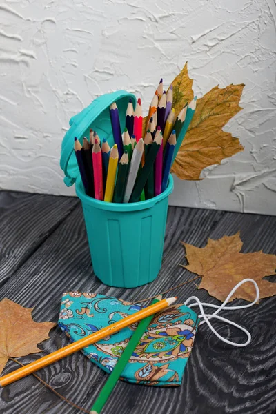 A pencil case in the form of a trash can. It contains colored pencils. Nearby is a protective face mask for the period of a pandemic. Dried maple leaves are added to the compositions.