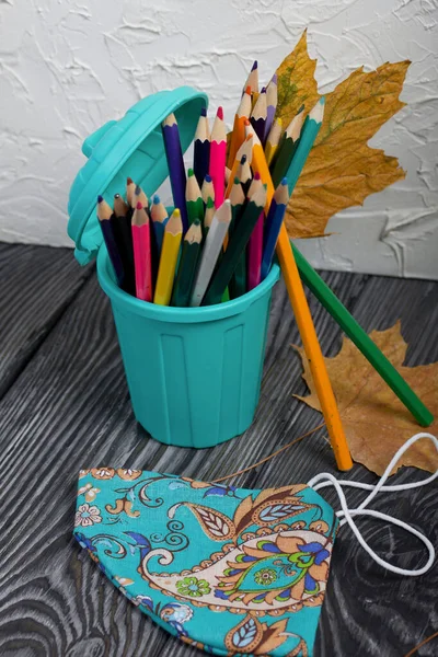 A pencil case in the form of a trash can. It contains colored pencils. Nearby is a protective face mask for the period of a pandemic. Dried maple leaves are added to the compositions.
