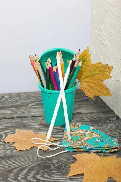 A pencil case in the form of a trash can. It contains colored pencils. Nearby is a protective face mask for the period of a pandemic. Dried maple leaves are added to the compositions.