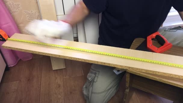 Um homem mede um tabuleiro com uma fita métrica. Marca a linha de corte com um lápis. Fazendo móveis em casa . — Vídeo de Stock