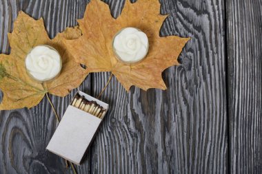 Çiçek şeklinde iki dekoratif mum. Yanlarında bir kutu kibrit ve kurumuş akçaağaç yaprakları var. Boyalı tahtalarda.