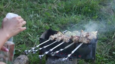 Şiş kebap ızgarada pişiyor. Bir kadın sebze ve et doğruyor. Kebap 'a su serper. Duman kömürlerden yayılıyor.