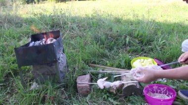 Yanan odunlu metal brazier. Ateş ve duman görünür. Adamın biri tavuk etini şişlere bağlıyor. Barbekünün yanındaki yerler.