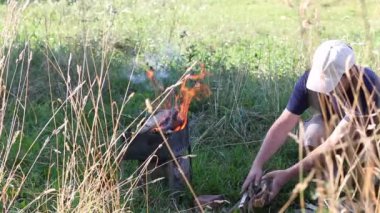 Yanan odunlu metal brazier. Ateş ve duman görünür. Adam onun yanına odun yığıyor. Uzun kuru otlar yetişir..