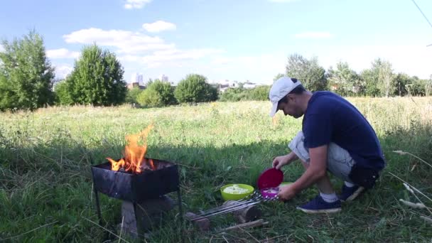 Металевий мангал з палаючою деревиною. Вогонь і дим видно. Чоловік перевіряє контейнери з м'ясом і овочами, оглядає шампури . — стокове відео