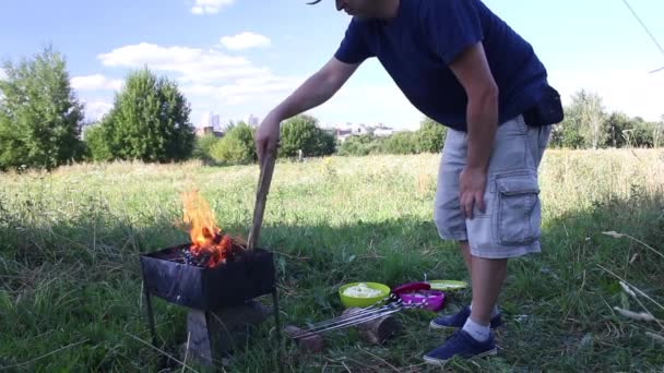 Металевий мангал з палаючою деревиною. Вогонь і дим видно. Чоловік контролює вогонь. На траві поблизу є контейнери з м'ясом та овочами . — стокове відео