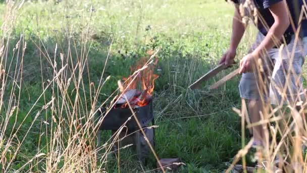 Kohlenbecken aus Metall mit brennendem Holz. Feuer und Rauch sind zu sehen. Der Mann richtet das brennende Holz auf. Hohes trockenes Gras wächst herum. — Stockvideo