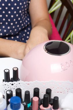 Bir kadın kendine manikür yaptırır. Verniği ultraviyole lambada kurutur. Şişedeki ojenin yanında..