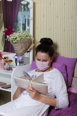 Tıbbi maskeli hasta bir kadın. Yatağa uzan. Fotoğraf albümüne bak. Yatağın yanında bir buket var. .